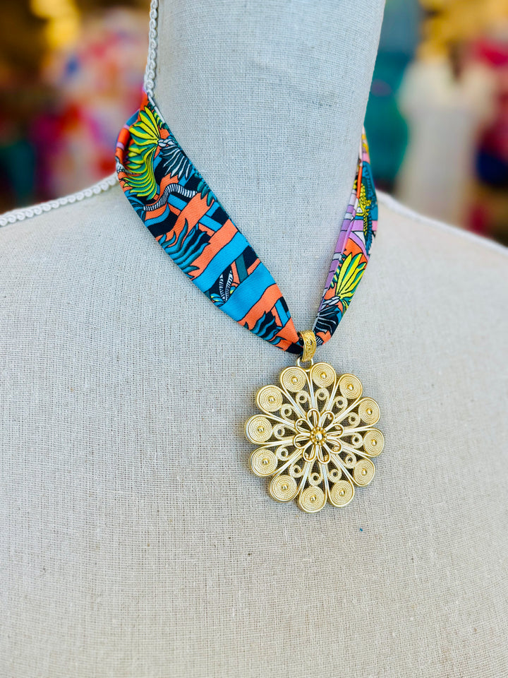 Tropical-inspired scarf necklace featuring a vibrant, multicolor fabric with jungle foliage patterns, accented by a shiny gold medallion charm at the center. Perfect for adding a bright, exotic flair to any outfit.