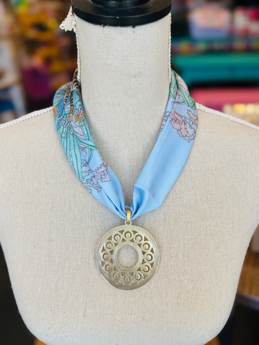 Blue printed scarf necklace with the words “lovely tiger,” featuring a gold cutout medallion charm; a playful and whimsical accessory perfect for adding a touch of charm and elegance to any outfit.