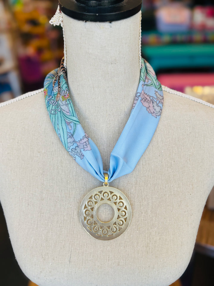 Blue printed scarf necklace with the words “lovely tiger,” featuring a gold cutout medallion charm; a playful and whimsical accessory perfect for adding a touch of charm and elegance to any outfit.