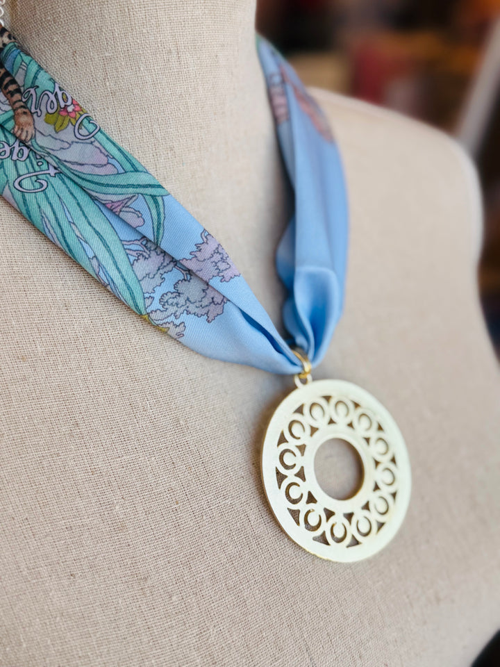 Blue printed scarf necklace with the words “lovely tiger,” featuring a gold cutout medallion charm; a playful and whimsical accessory perfect for adding a touch of charm and elegance to any outfit.