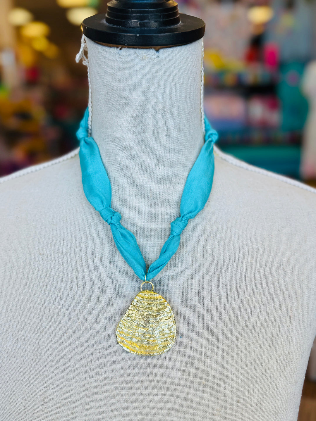Close-up image of a blue scarf-style necklace with a textured gold medallion charm, styled to evoke breezy coastal vibes. The soft aqua blue fabric contrasts elegantly with the bold gold pendant, creating a light and stylish accessory perfect for casual or chic outfits.