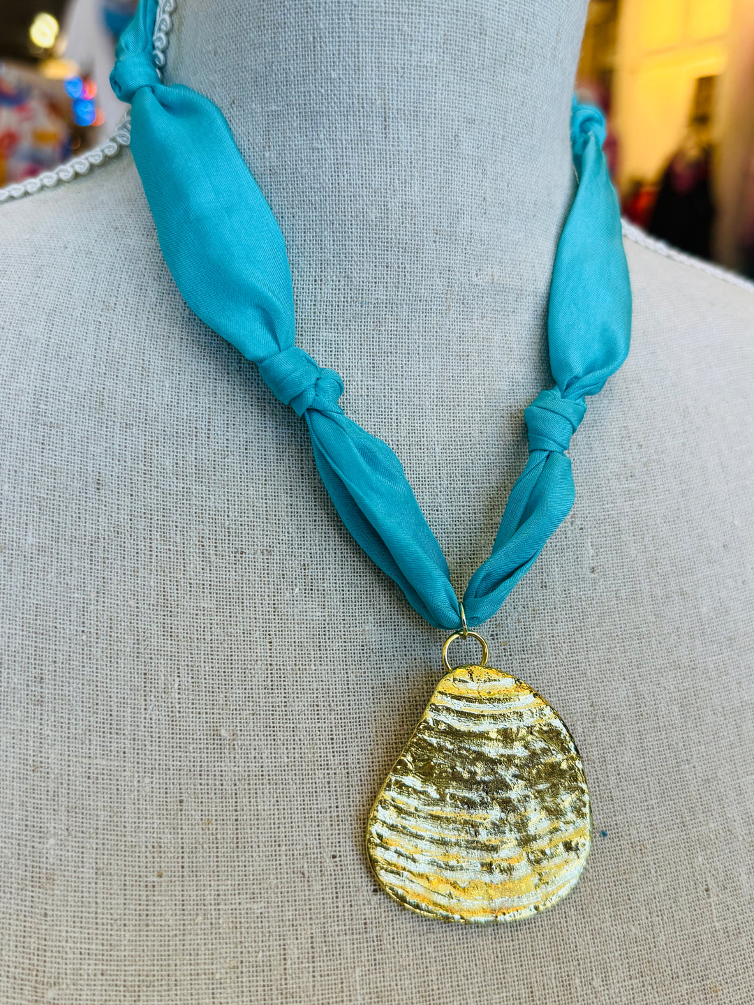 Close-up image of a blue scarf-style necklace with a textured gold medallion charm, styled to evoke breezy coastal vibes. The soft aqua blue fabric contrasts elegantly with the bold gold pendant, creating a light and stylish accessory perfect for casual or chic outfits.