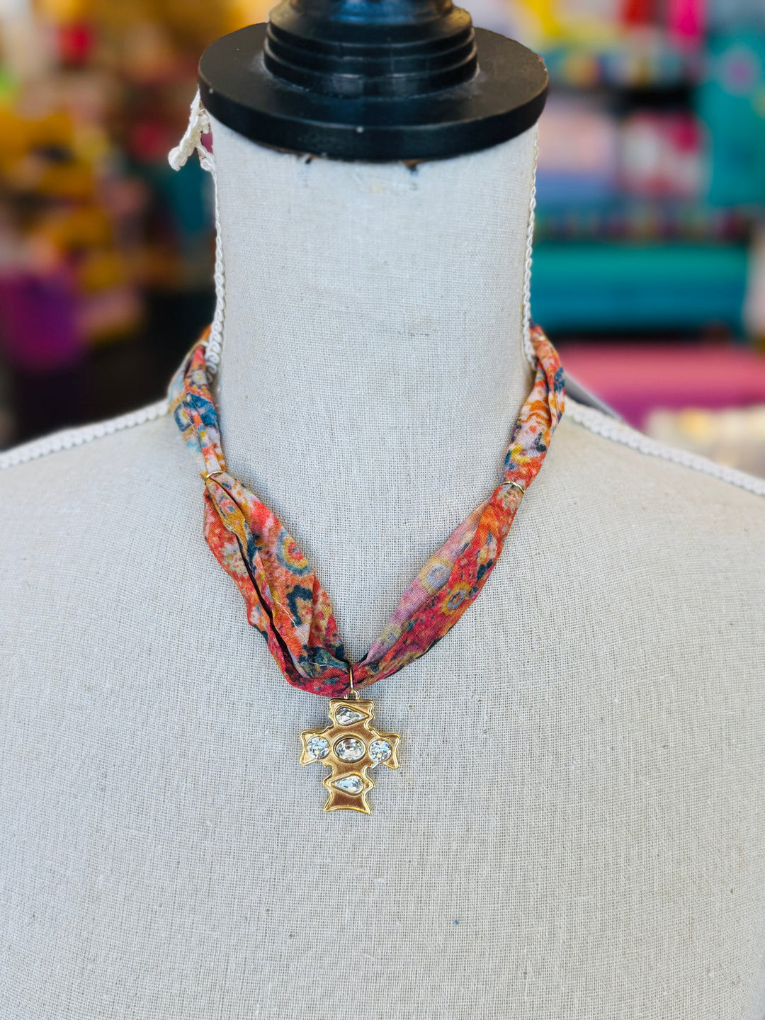 Scarf necklace featuring a richly patterned fabric band and a sculpted gold cross charm with sparkling crystal accents, styled to add vintage charm and elegance to any outfit.