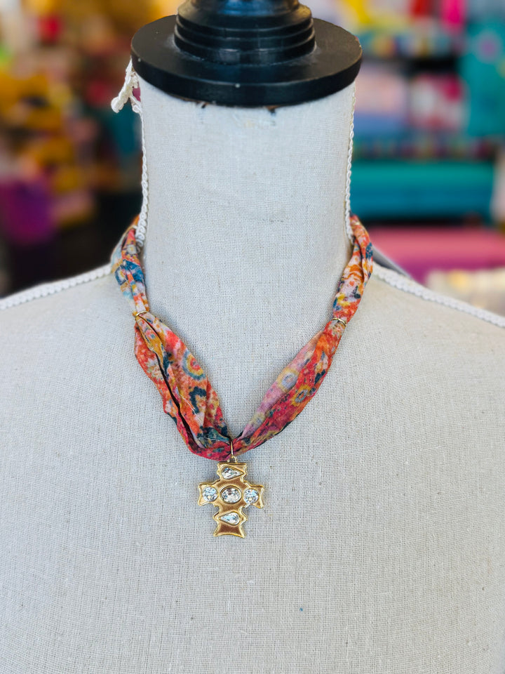 Scarf necklace featuring a richly patterned fabric band and a sculpted gold cross charm with sparkling crystal accents, styled to add vintage charm and elegance to any outfit.