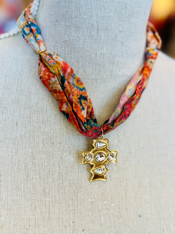 Scarf necklace featuring a richly patterned fabric band and a sculpted gold cross charm with sparkling crystal accents, styled to add vintage charm and elegance to any outfit.