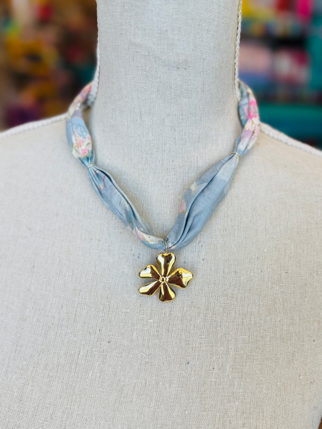 Close-up of a blue printed scarf necklace with soft pink accents, featuring a bold gold flower charm pendant, styled flat on a white surface for a romantic and feminine accessory look.