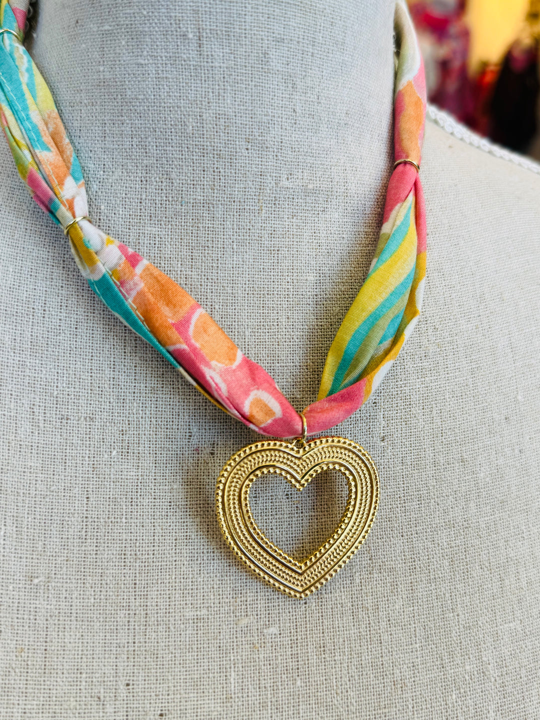 Colorful abstract print scarf necklace featuring a textured open gold heart charm, styled as a playful and lightweight accessory perfect for adding a bold pop of color and charm to any outfit.