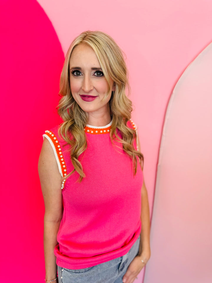 Pink sweater vest with orange and white striped neckline and armholes, worn with jeans; pearl detailing visible on the vest front.