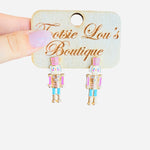 Pastel nutcracker earrings with pink hats, teal pants, gold sparkle trim, and cheerful faces.