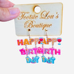 A pair of playful dangle earrings featuring colorful charms, including letter beads that spell out "Happy Birthday," a miniature party hat, and a small wine glass.