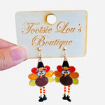 A pair of dangle earrings shaped like turkeys. The turkeys have colorful enamel tail feathers, are wearing small black pilgrim hats, and have little legs that dangle.