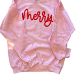 Soft pink sweatshirt with bold red “Merry” script lettering in festive holiday style.