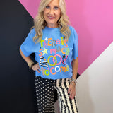 Blue graphic tee with colorful Good To Come lettering, stripes, checks, stars, and playful patterns.