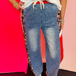 Denim jogger pants with rhinestone embellishments, red side stripes, and leopard print panels for a bold boutique fashion look.