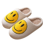 Cream plush slippers with oversized yellow smiley face design, soft and cozy for lounging.