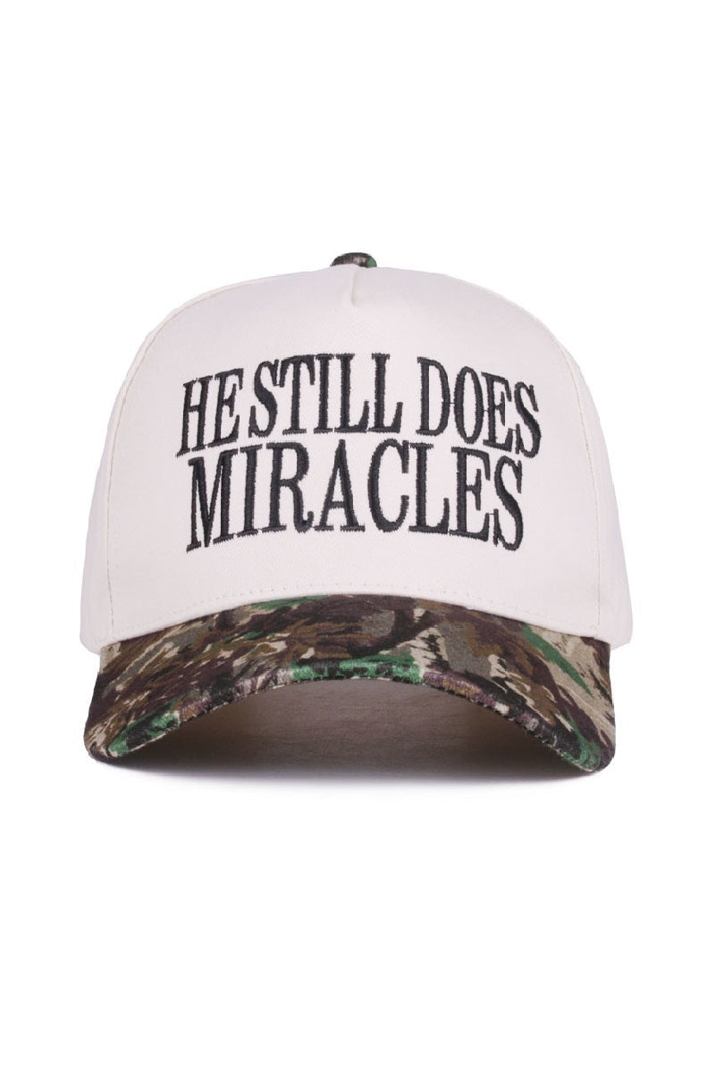 Zenana White cap with black “He Still Does Miracles” embroidery and camo bill, adjustable strap.