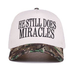 Zenana White cap with black “He Still Does Miracles” embroidery and camo bill, adjustable strap.