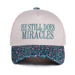 Zenana White cap with turquoise “He Still Does Miracles” embroidery and cougar-print bill, adjustable fit.