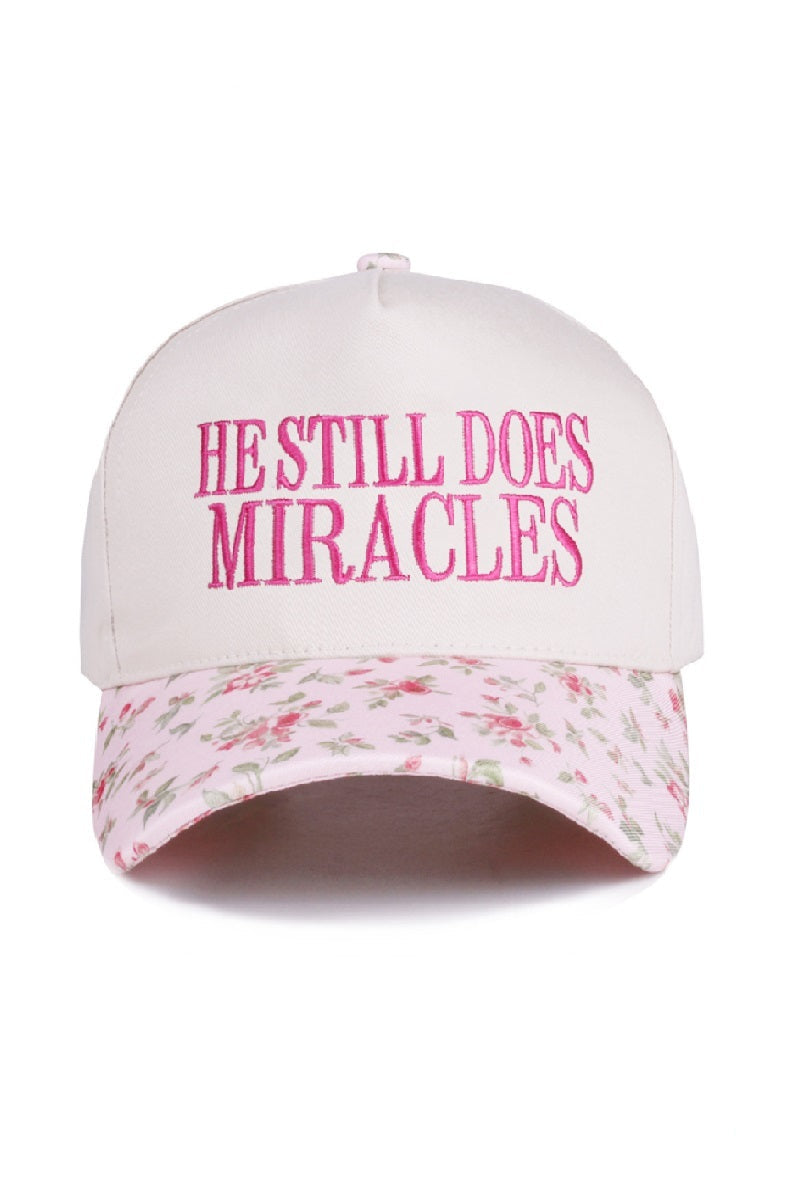 Zenana White cap with pink “He Still Does Miracles” embroidery and floral-print bill, adjustable strap.