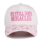 Zenana White cap with pink “He Still Does Miracles” embroidery and floral-print bill, adjustable strap.
