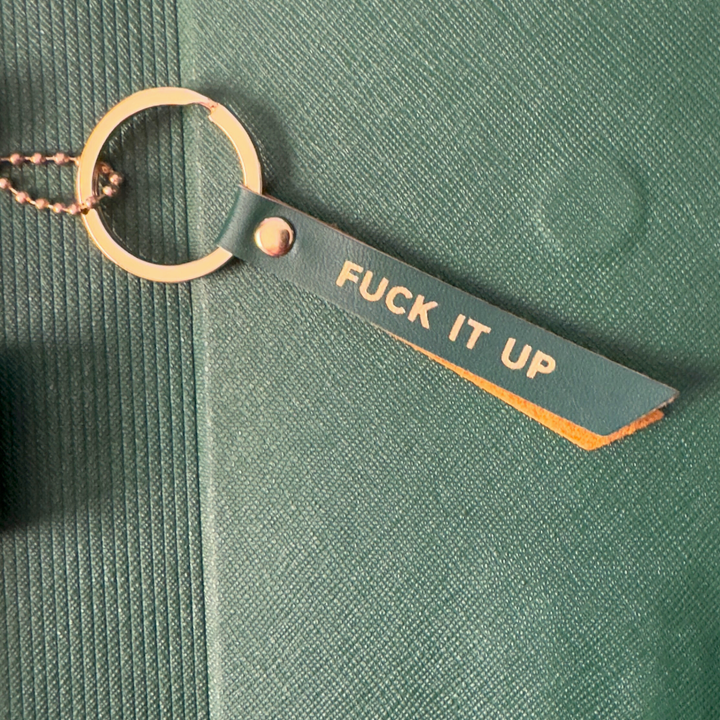 Keychain with 'FUCK IT UP' tag on a green textured surface