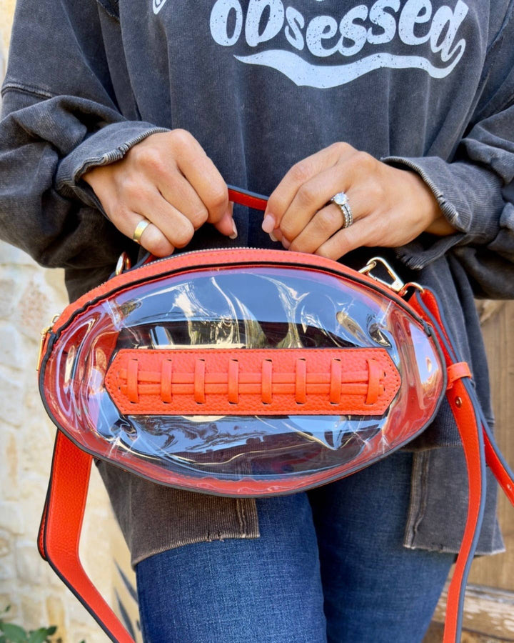 Clear football-shaped crossbody bag with orange trim, gold hardware, top handle, and adjustable strap, displayed to show shape and transparency.