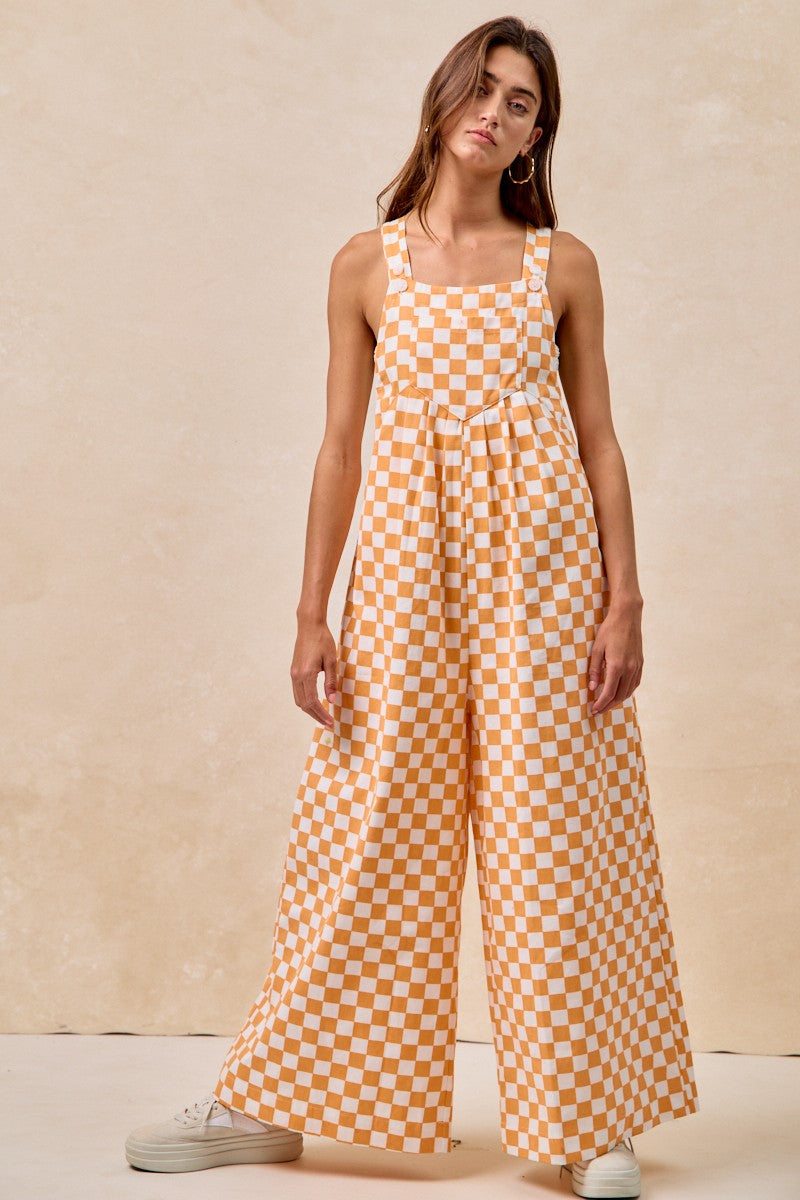 A pair of overalls with an orange and white checkerboard plaid print. The overalls have adjustable straps, front pleats, and a wide-leg silhouette.