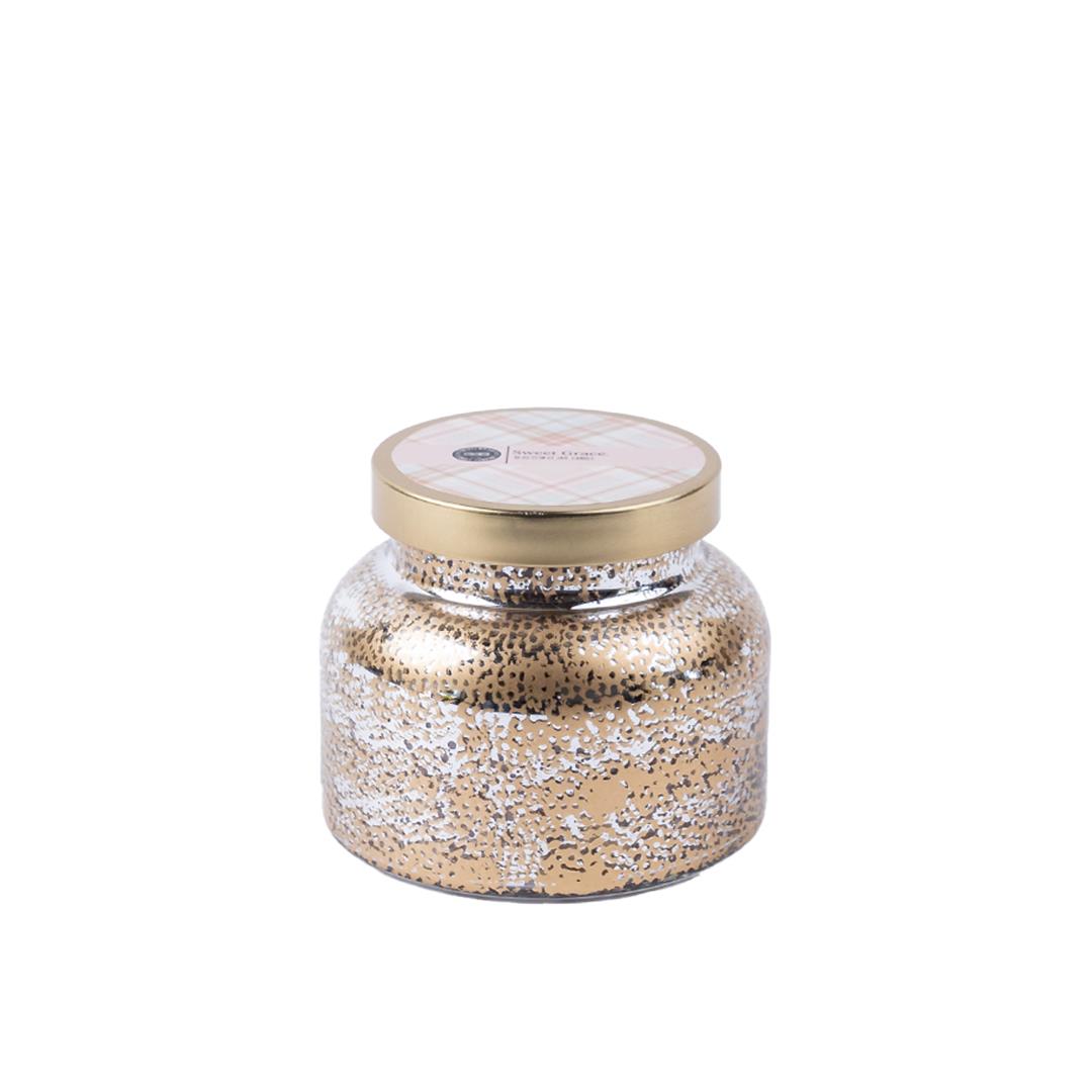 A clear jar candle with a gold lid, decorated with a textured design and a label in the center.