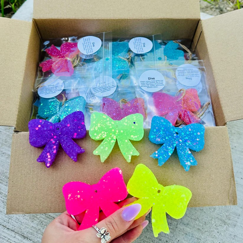 A car air freshener shaped like a small, glittery bow, shown in a vibrant, assorted color.